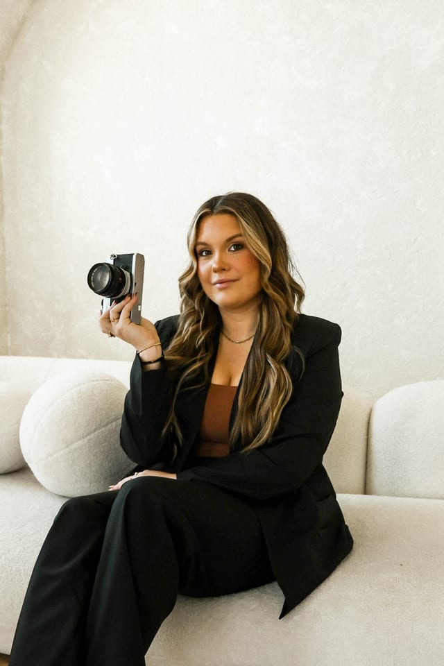 Madi Atkisson holding a vintage camera, seated on a cream couch in a black suit