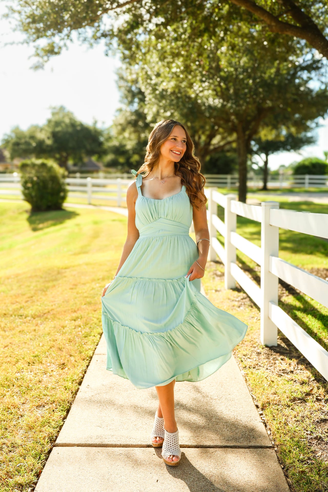 Senior in mint dress walking a sunlit path beside a white fence